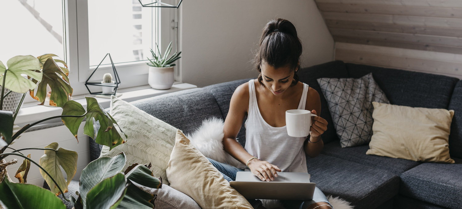 [Featured image] A person holds a cup of coffee while using their laptop on the couch.