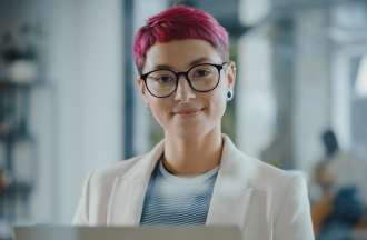 [Featured Image]:  A female, with red hair, wearing glasses, a white jacket and a blue and white top is sitting at her desk in front of her computer, performing her duties as a Health Care Data Analyst. 
