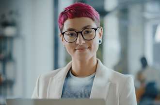 [Featured Image]:  A female, with red hair, wearing glasses, a white jacket and a blue and white top is sitting at her desk in front of her computer, performing her duties as a Health Care Data Analyst. 