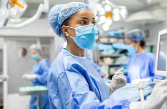 [Featured image] A nurse anesthetist wearing surgical scrubs and a mask checks a monitor in an operating room of a hospital.