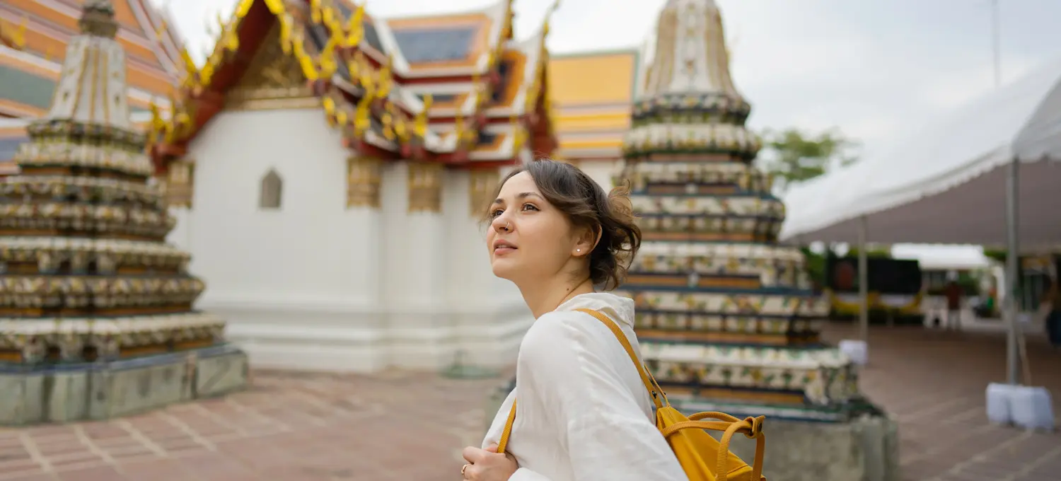 [Featured Image] Young person on a gap year trip exploring Wat Pho in Bangkok while wearing a white dress and yellow backpack.