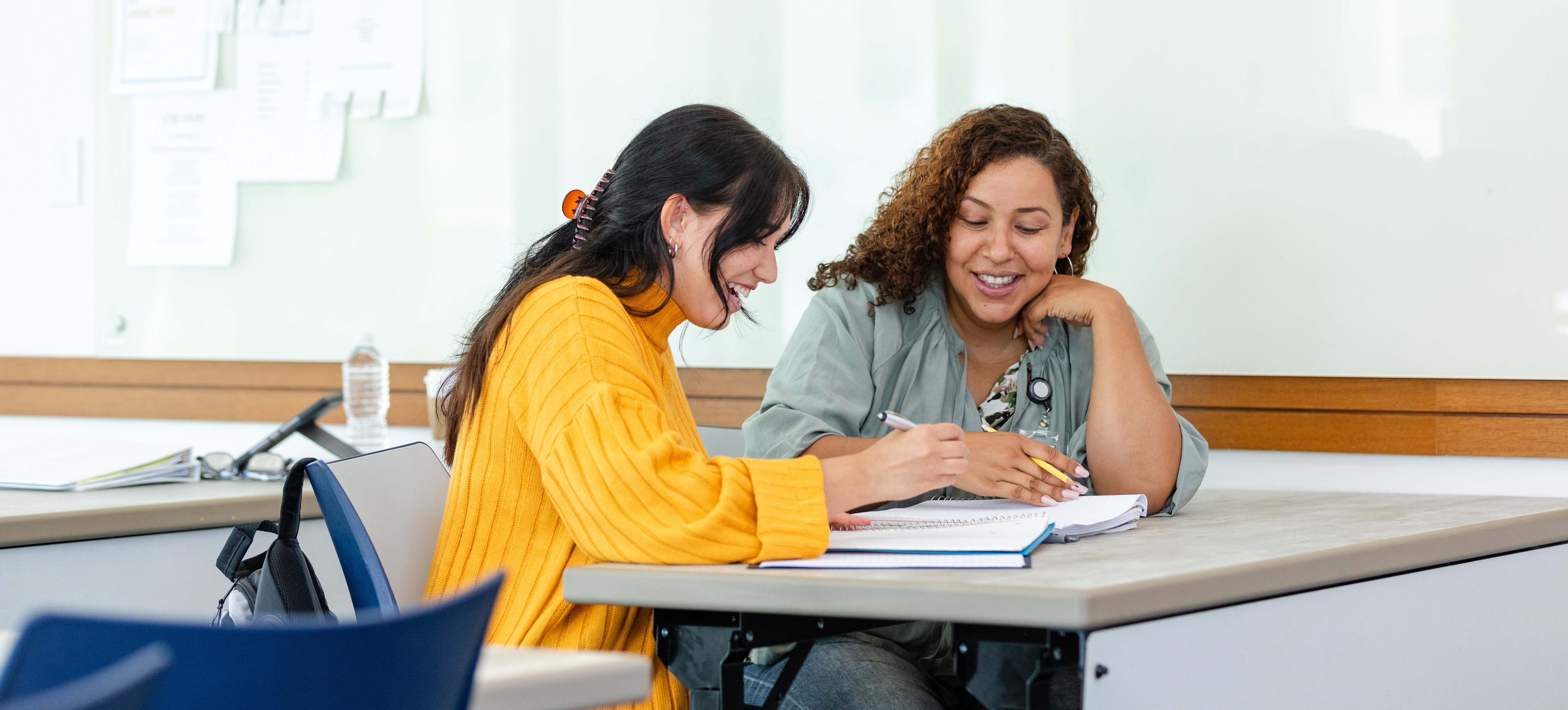 [Featured Image] A female teacher sits at a desk with a female student in a classroom. The teacher is explaining how to revise.