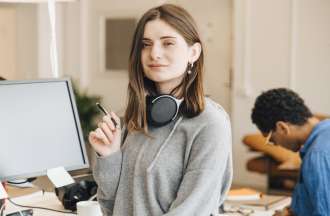 [Featured image] A young white woman in a gray long-sleeved top and over-the-ear headphones around her neck stands in front of a desktop computer. 