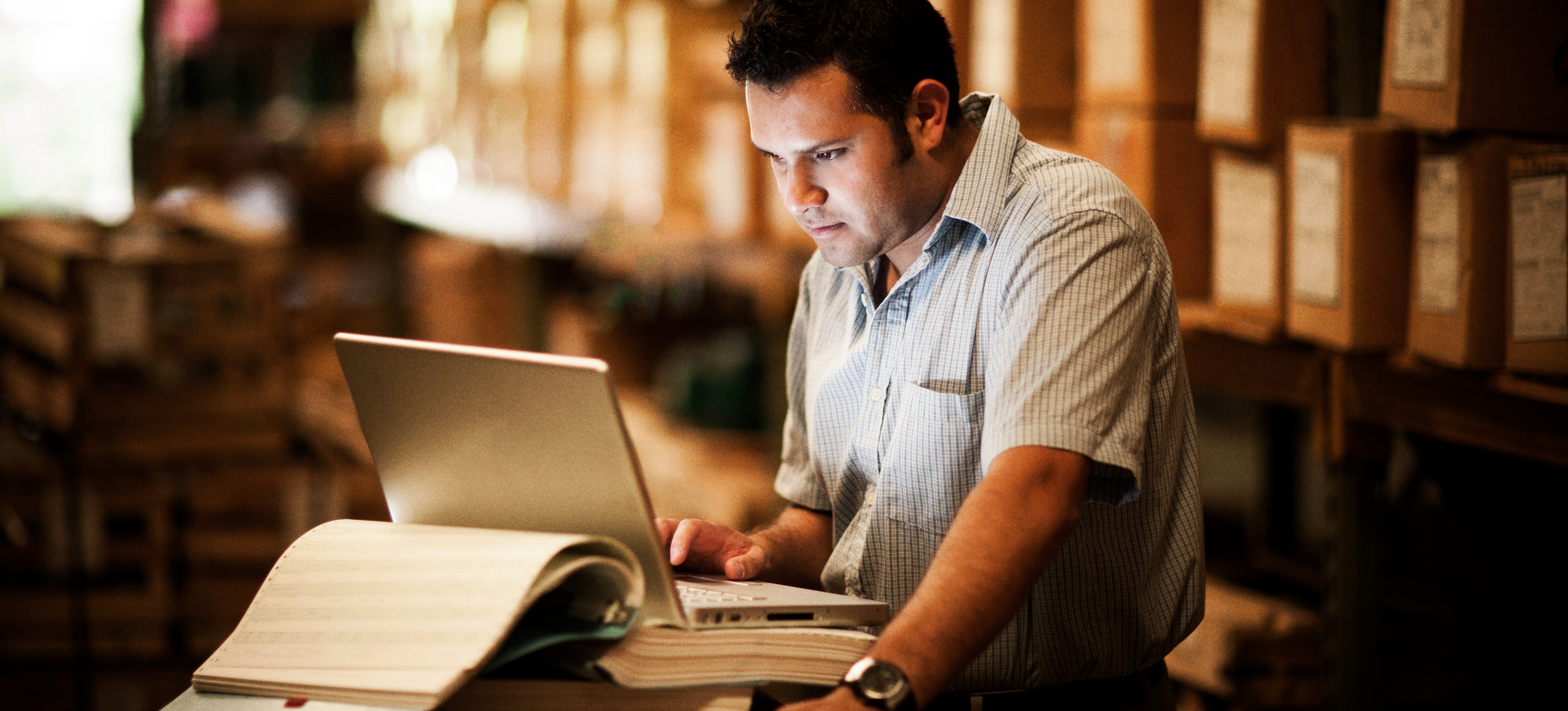 [Featured Image] A warehouse manager stands in front of shelves packed with products as they work with the Google Sheets checkbox feature on their laptop.