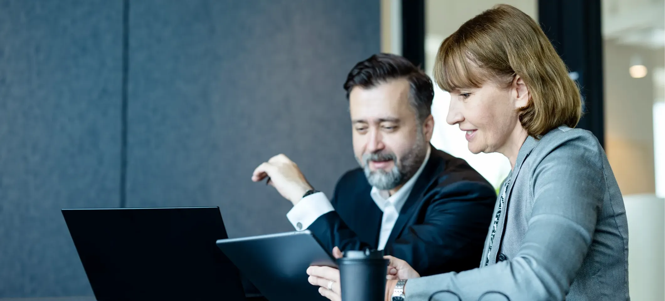 [Featured image]: Two people sitting in a conference room look at a tablet and laptop as they discuss accounting topics.
