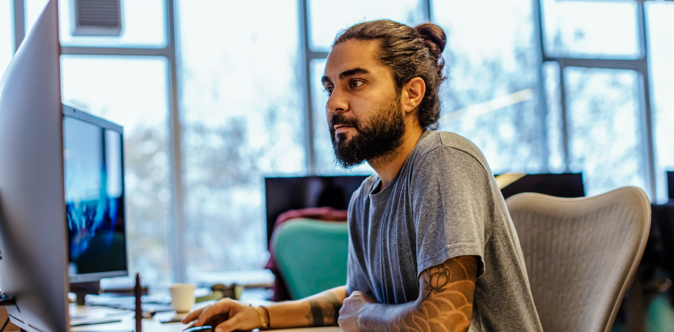 [Featured image] A tattooed computer technician works at a dual-screen workstation in an office with windows behind them. 
