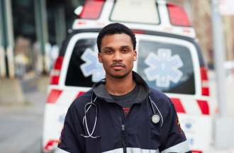 [Featured Image]: A man with short black hair and a mustache. He is wearing a black and white jacket. He has a stethoscope around his neck. He is standing in front of an ambulance. 