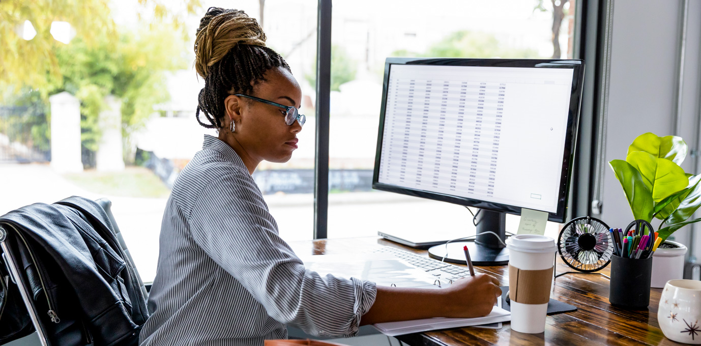 [Featured image] A person in glasses and a striped shirt sits in front of a computer, searching for business degree jobs while writing on a notepad.