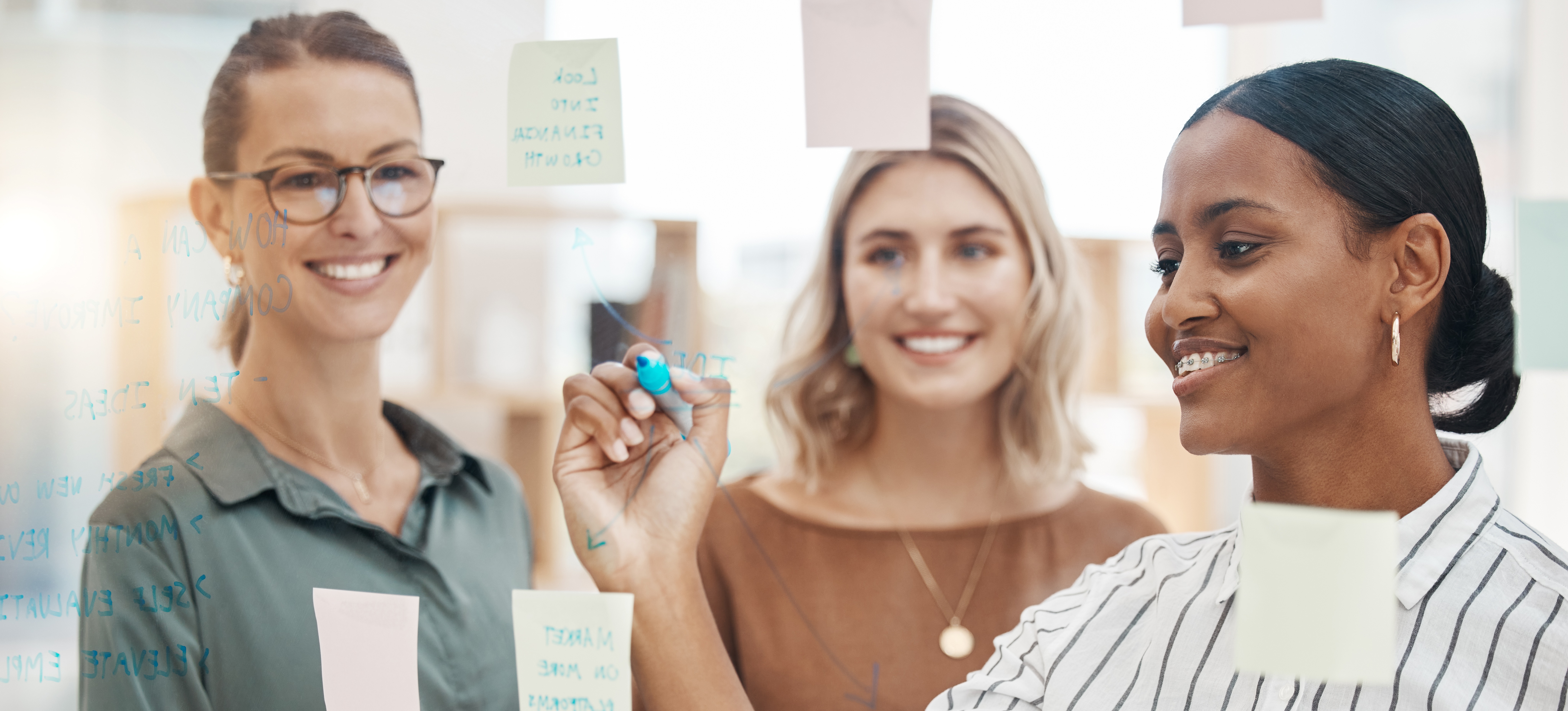 [Featured Image] A woman working for a market research analyst salary discusses her research with two colleagues as she writes on a transparent wipe board with a blue pen.
