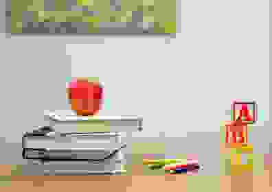 Desk with three books stacked and an apple on top of them, next to colored pencils and blocks stacked with the letters "A", "B", "C". 