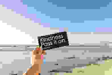A photo taken from the first person pov, showing someone holding out their arm holding a sticker that says kindness, pass it on.
