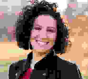 woman smiling with short dark curly hair, red blouse, and black buttoned jacket