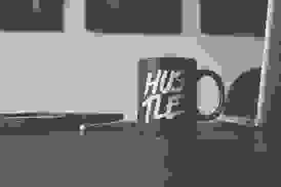A black mug with the word "hustle" printed on it sits on a brown office table