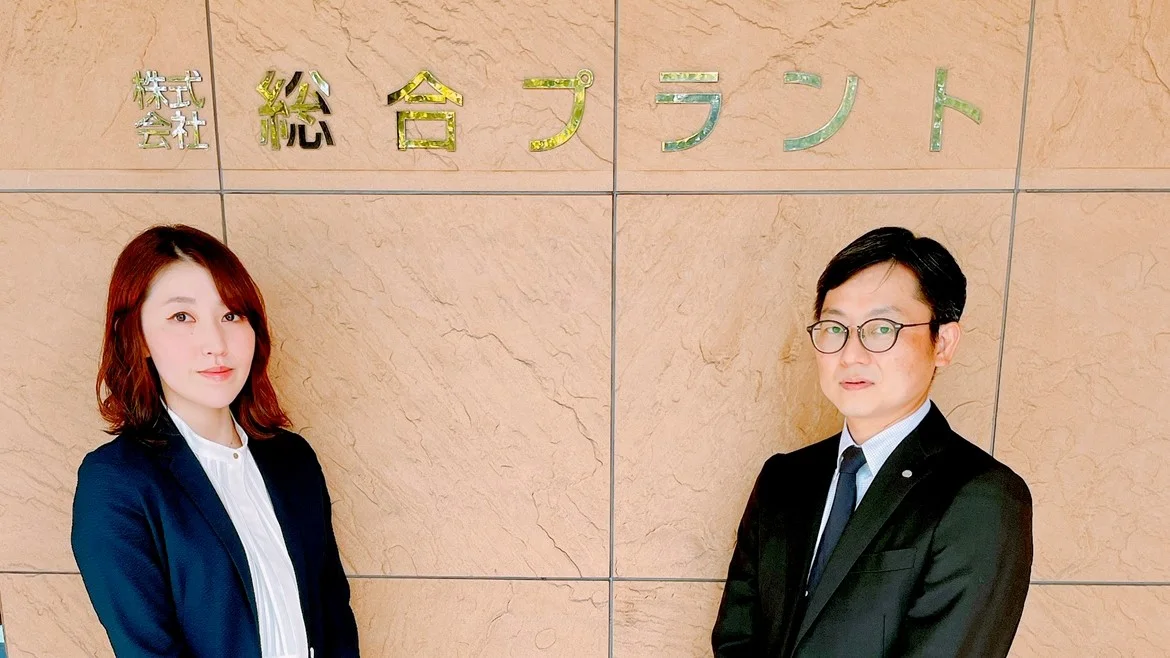 株式会社総合プラントの社名サイン前に立つ取材対象者2名