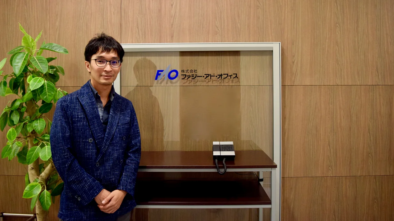 株式会社ファジー・アド・オフィスの社名プレート前に立つ取材対象者