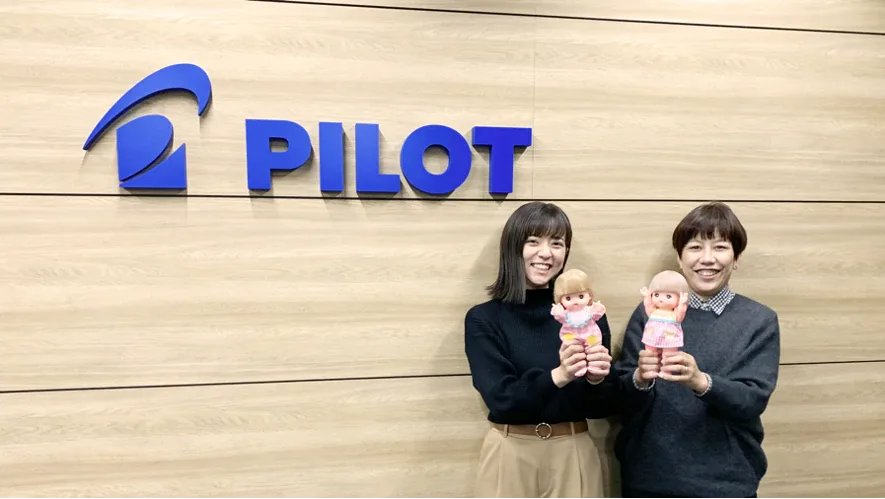PILOTのロゴが掲げられた壁の前で、メルちゃん人形を持つ取材対象者2名