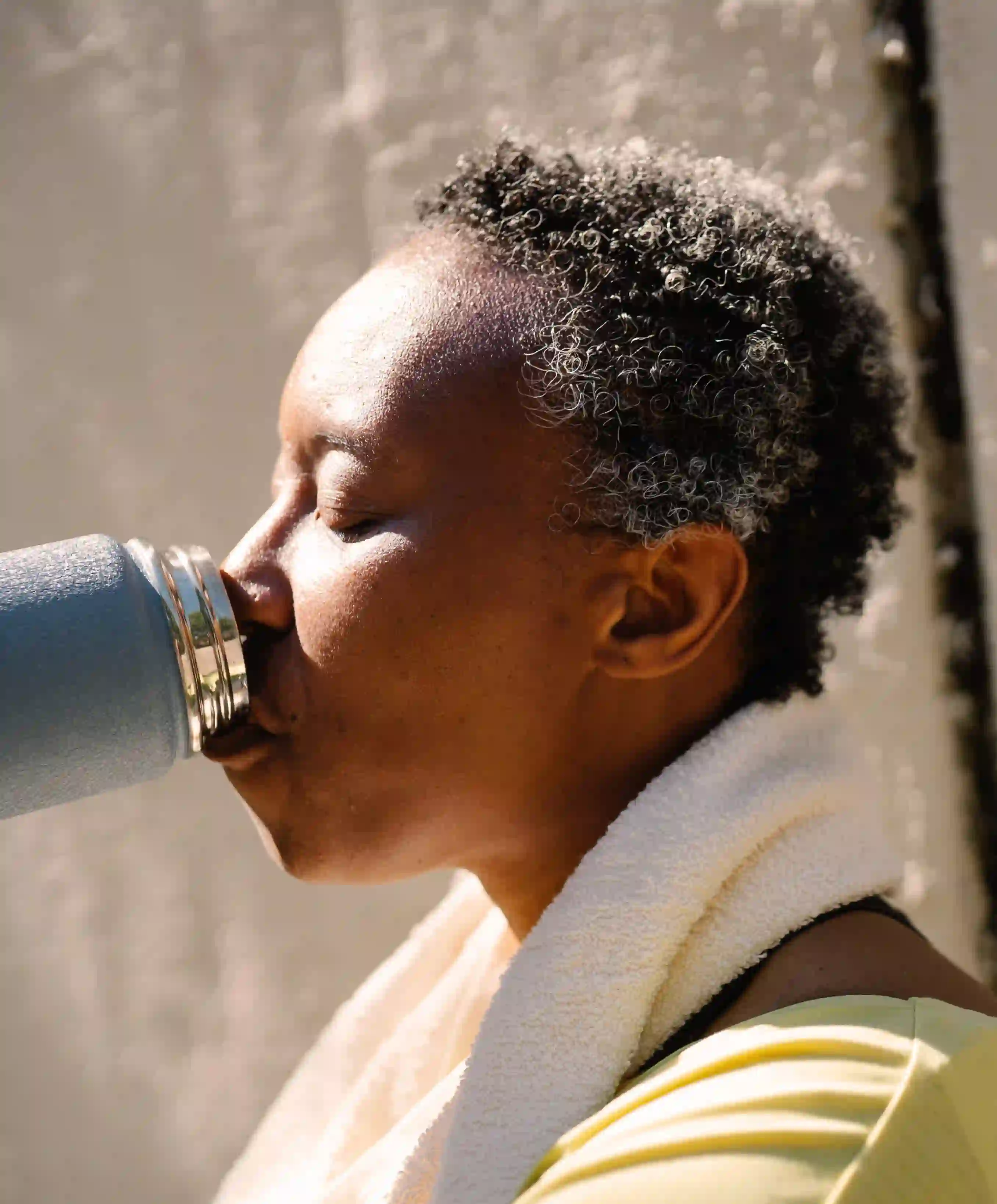A woman with a white towel around her neck drinking water from a thermos.