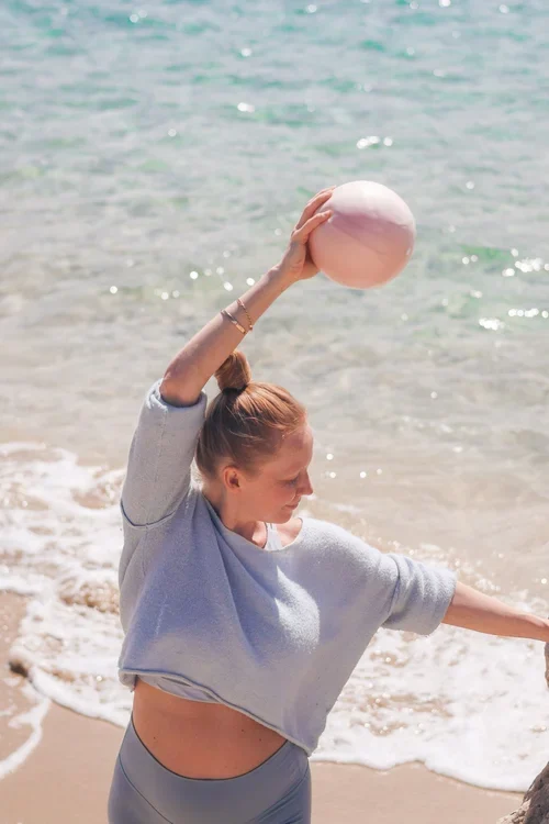 Person står vid strandkanten och sträcker en arm uppåt medan en träningsboll hålls över huvudet
