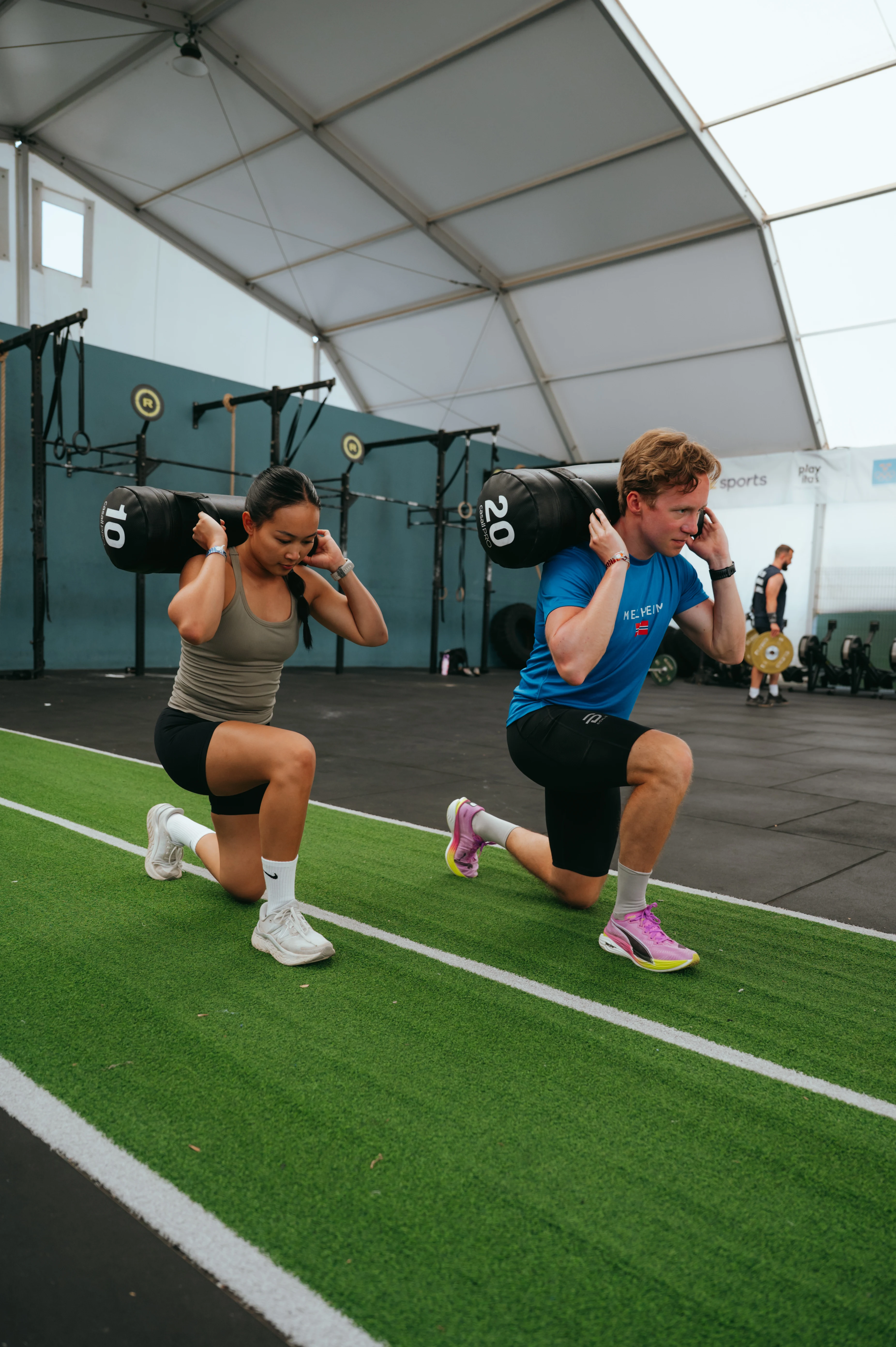 To personer utfører knebøy med vektsekker på skuldrene i et treningsstudio. De står på en grønn matte, og det er flere treningsapparater i bakgrunnen.