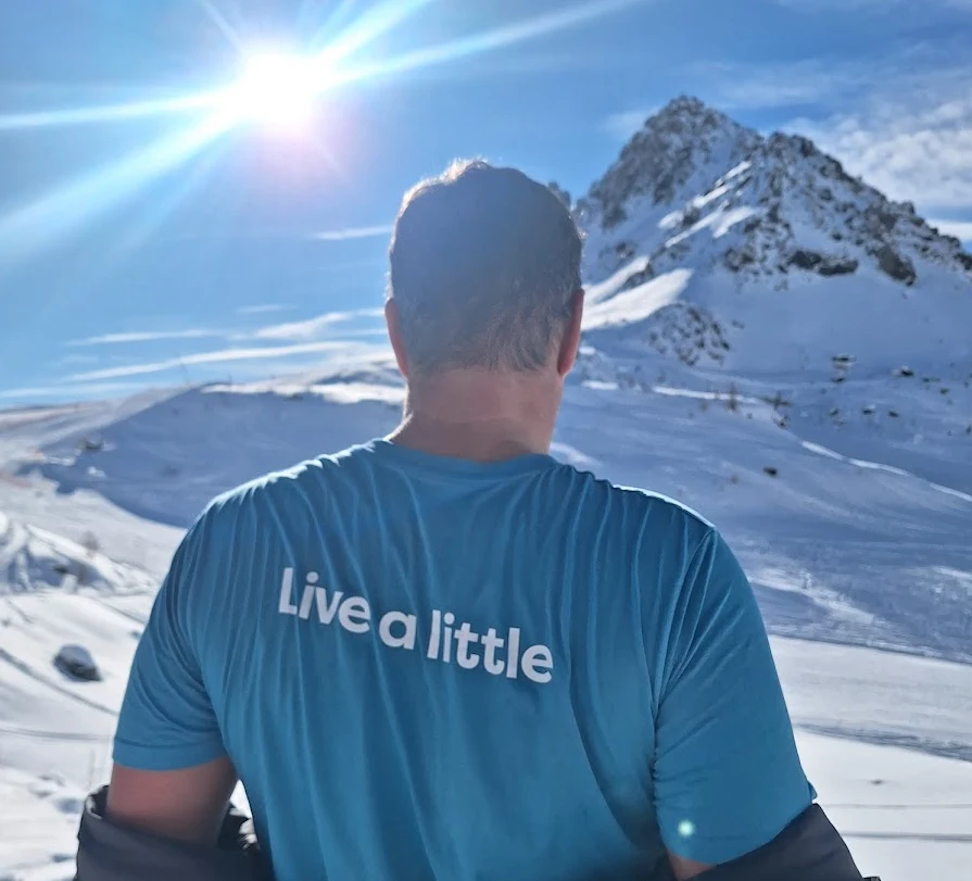 Person står i alperne med ryggen til i solskin med blå t-shirt hvor står der Live a Little.