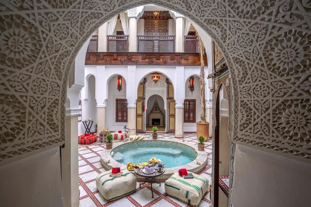 Traditionell innergård på Riad Safran et Cannelle & Spa i Marrakech med rund pool, marockanska sittplatser, vita valvbågar och dekorativa detaljer i trä och sten.
