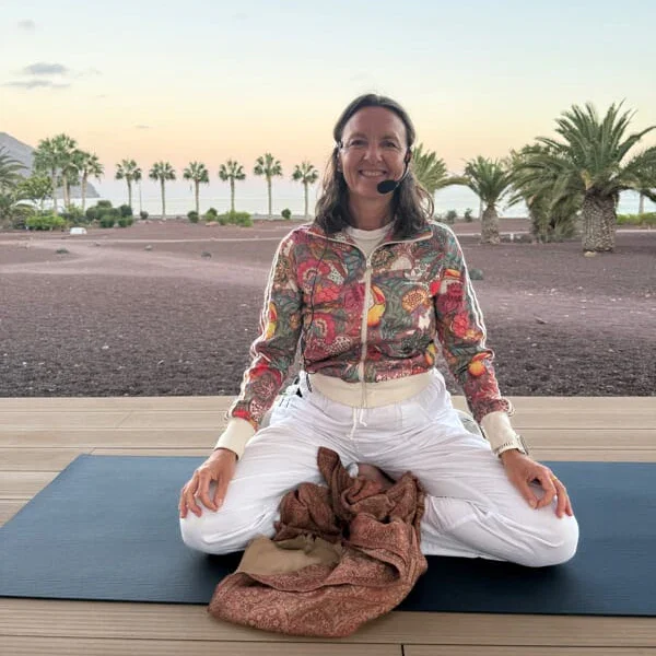 Profilbilde Ida Marie Kramer som sitter i Lotusstilling på en yogamatte med palmer og hav i bakgrunnen.