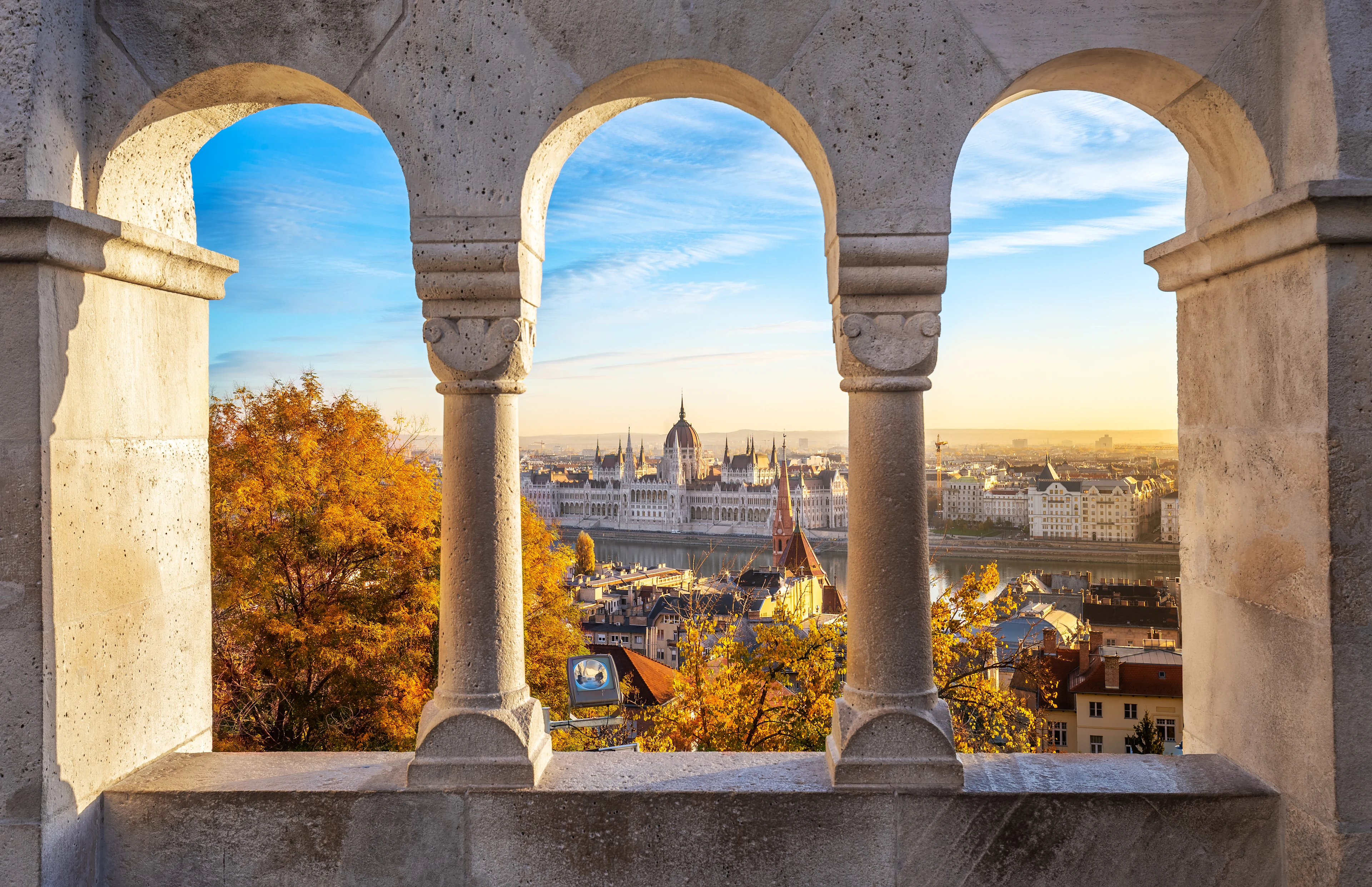 Utsikt över Budapest genom tre stenbågar, med parlamentsbyggnaden och Donau synliga i morgon- eller kvällsljus samt höstfärgade träd i förgrunden