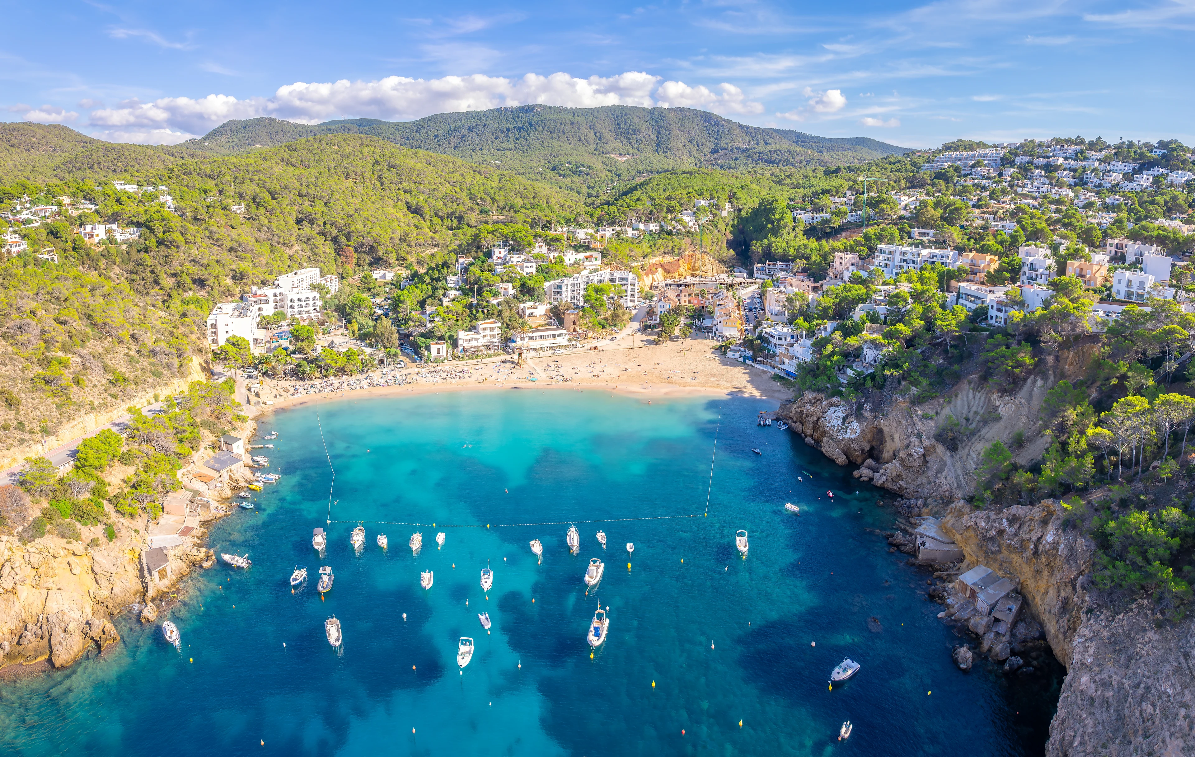 Flygfoto över Cala Vadella på Ibiza, med turkost vatten, segelbåtar och sandstrand