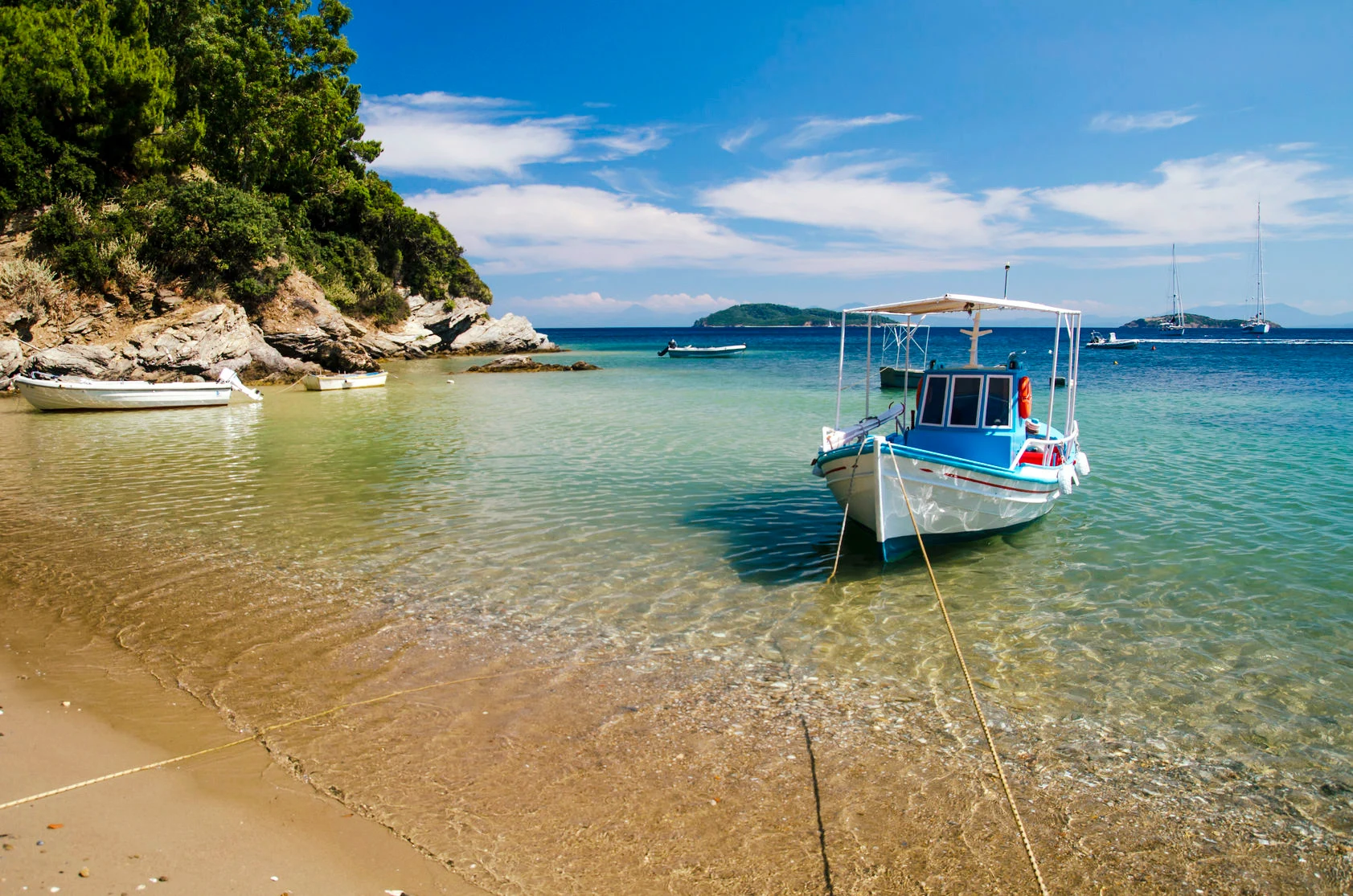 Liten vit och blå fiskebåt förtöjd vid en sandstrand med klart, turkost vatten, omgiven av gröna klippor och lugnt hav under en klarblå himmel på Skiathos i Grekland.