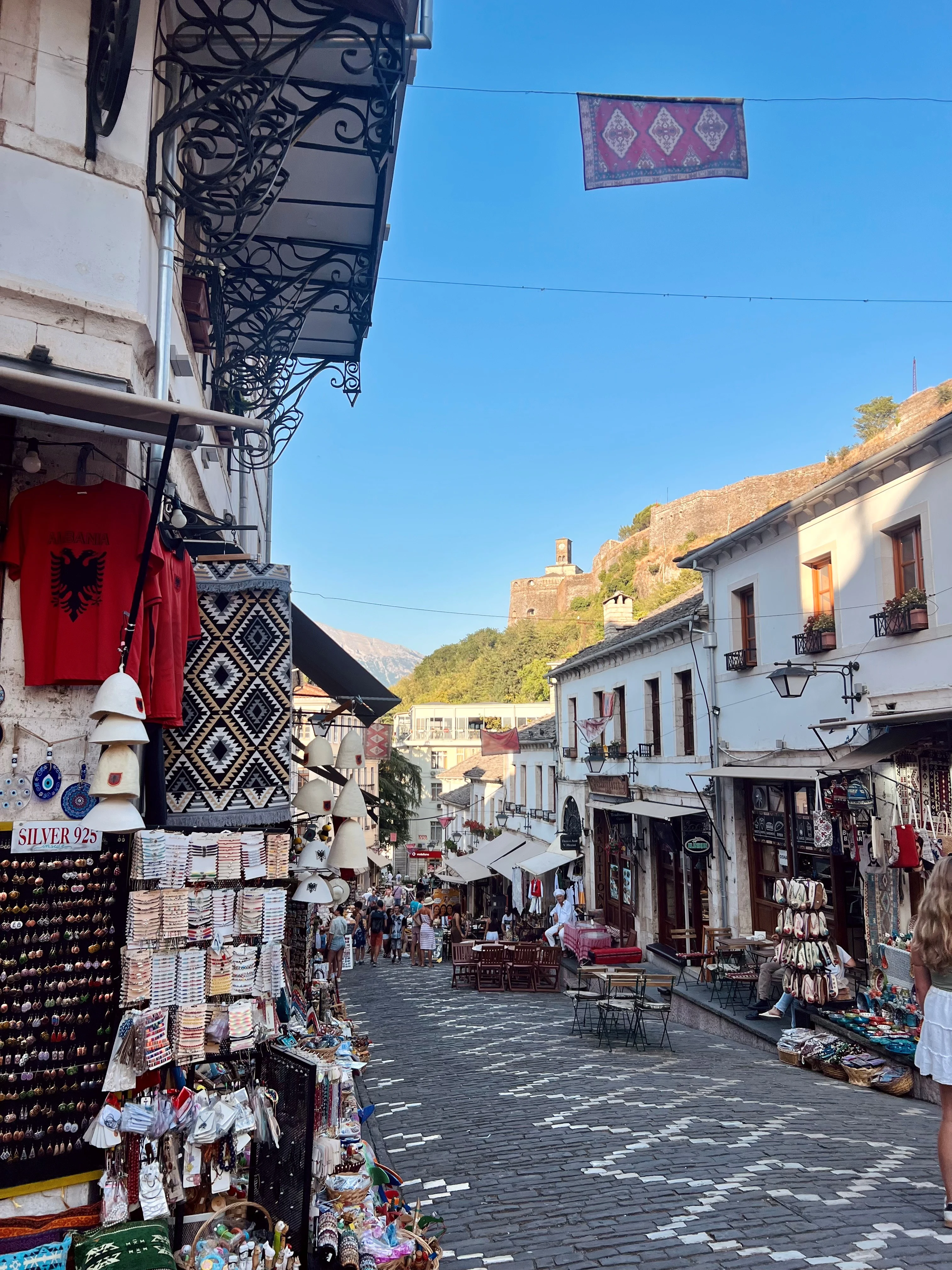 Gata i Gjirokaster med hus som kantas av butiker med ett berg och slott i bakgrunden. 