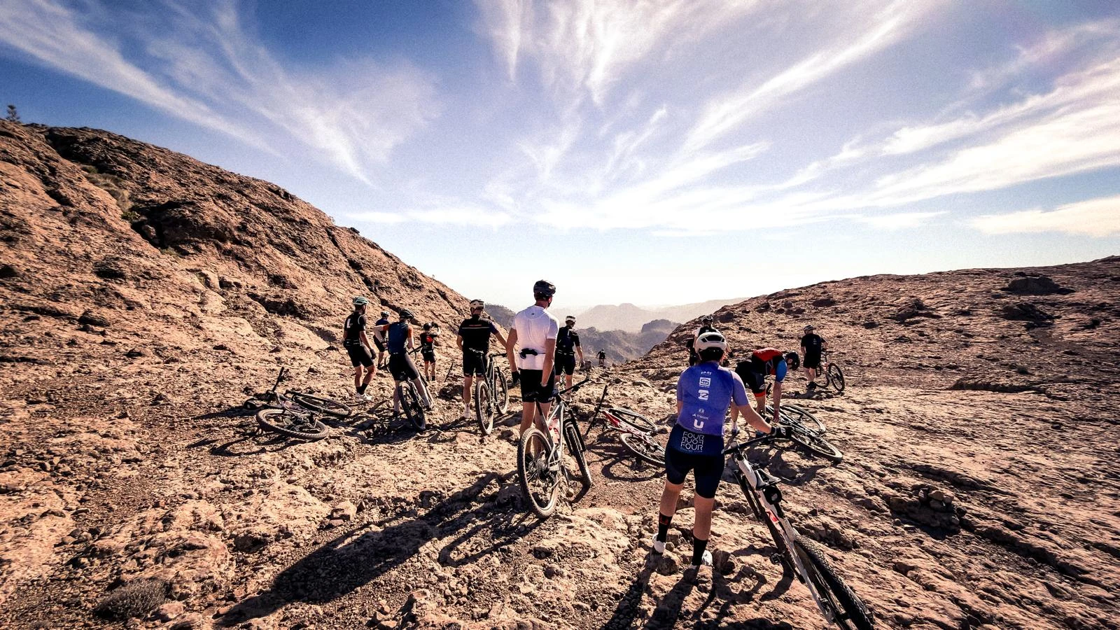 En grupp cyklister står med sina mountainbikes på ett öppet, stenigt bergsområde med vid utsikt över dalar och klippor under en klar himmel