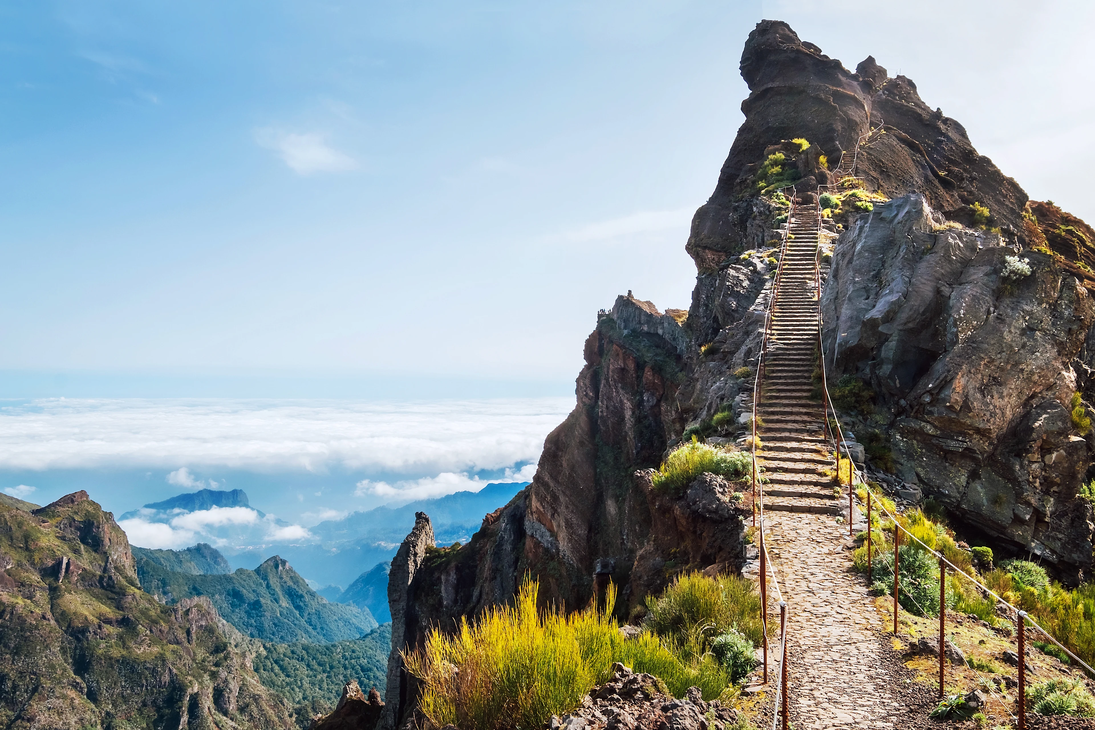 Tursti med rekkverk som fører opp til en spiss fjelltopp høyt oppe i fjellene på Madeira. I bakgrunnen ses hvite skyer, blå himmel og mer magisk Madeira natur.  