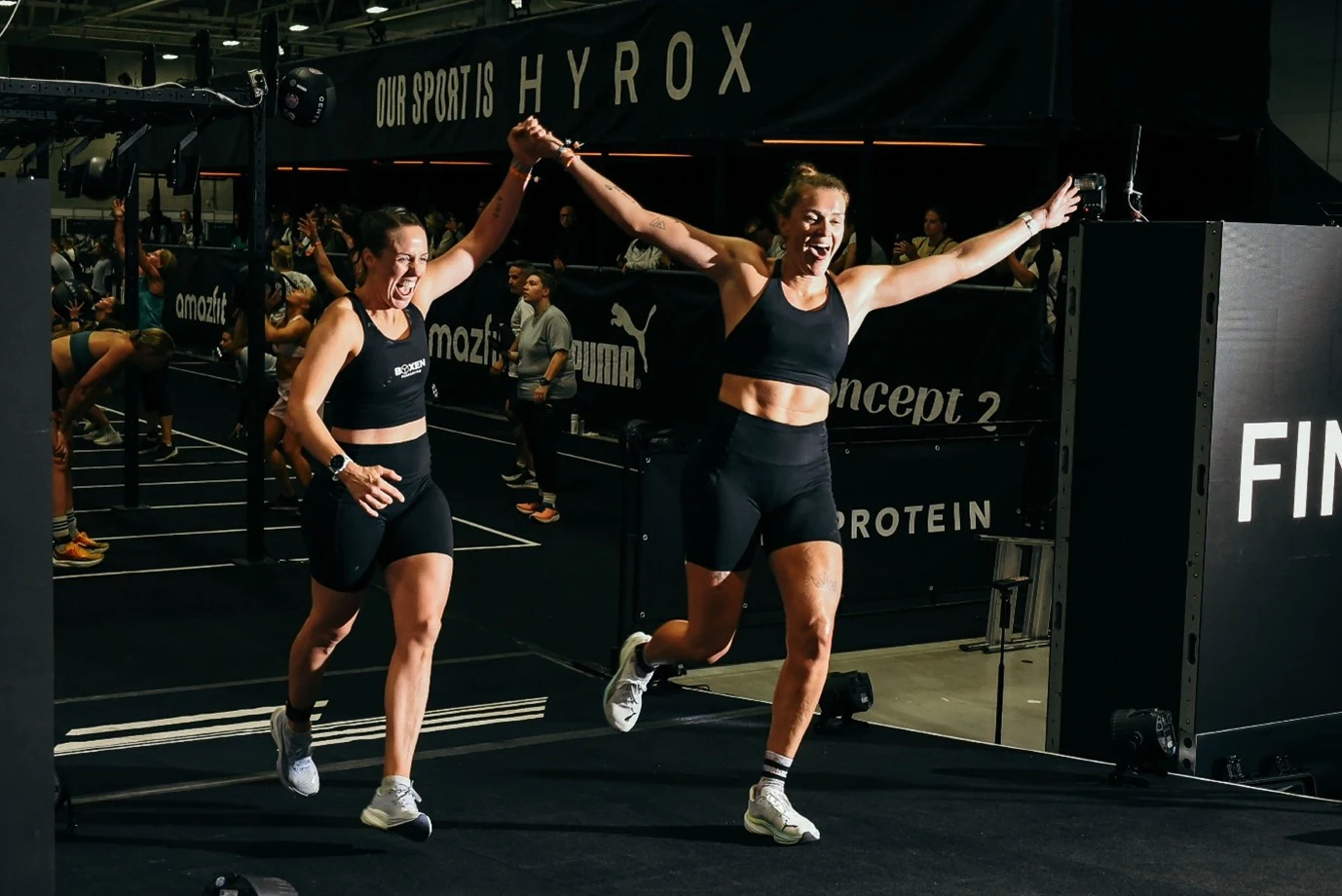 To damer ikledd sorte treningsshorts og sorte treningstopper som løper inne på et treningsrtudio mens de holder hendene i været.