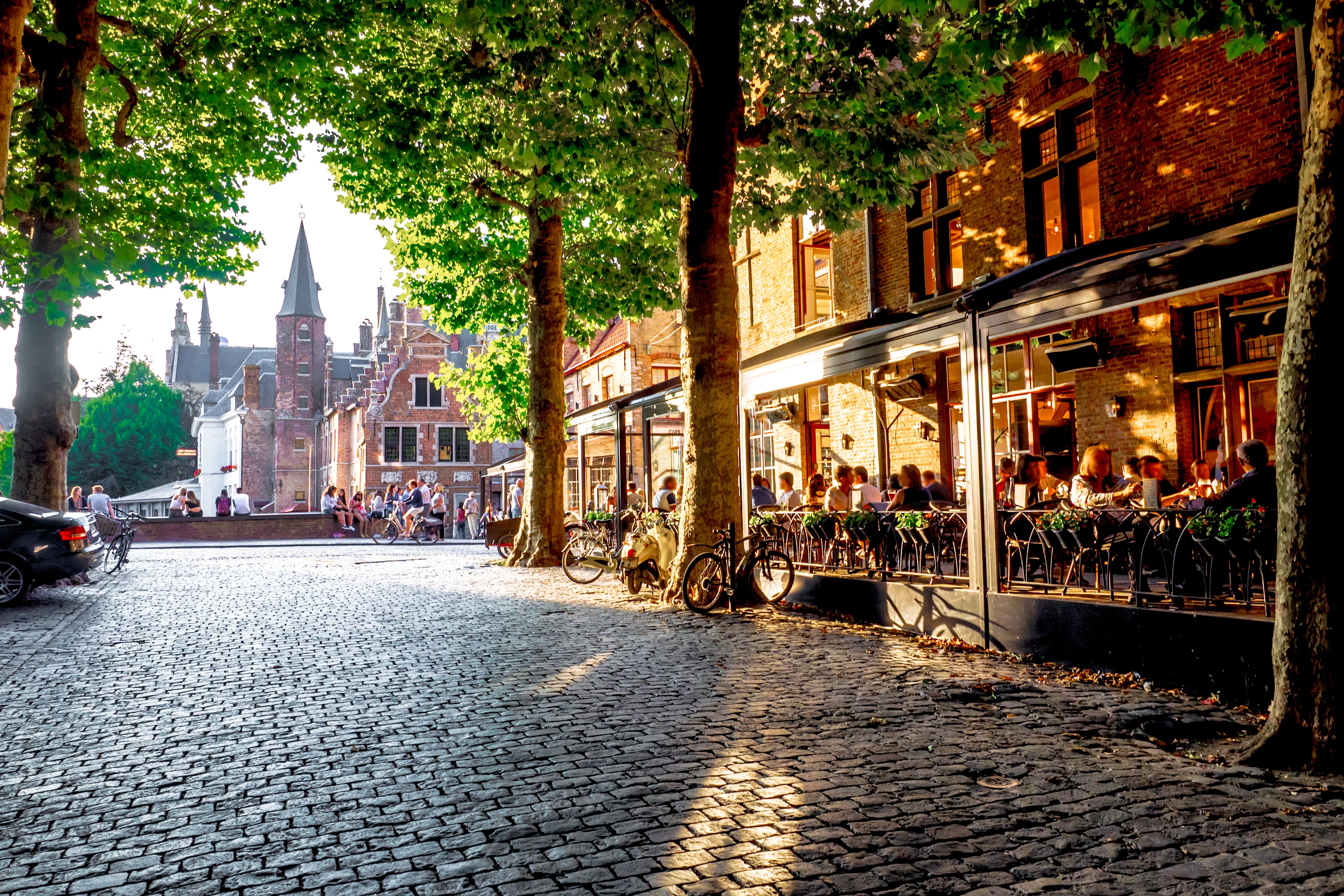 Kullerstenstorg med uteserveringar, träd, cyklar och historiska byggnader i stadsmiljö.