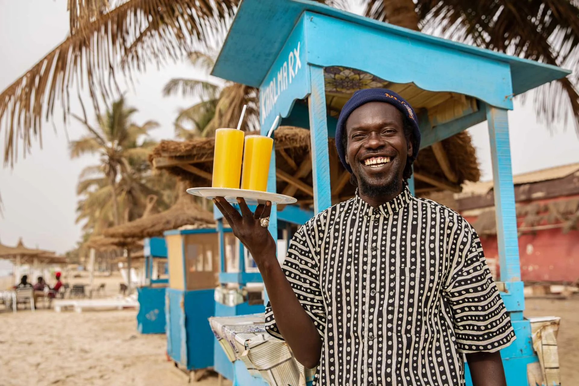 En man ler mot kameran, står på stranden, med en bricka i handen. På brickan står det 2 glas apelsinjuice.