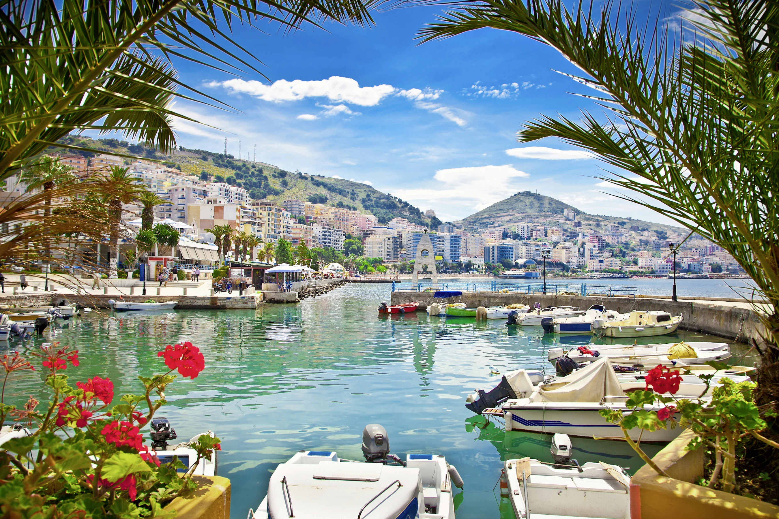 havnen i Saranda med havnepromenaden og husene i bakgrunnen. Motorbåter, palmer og blomster 