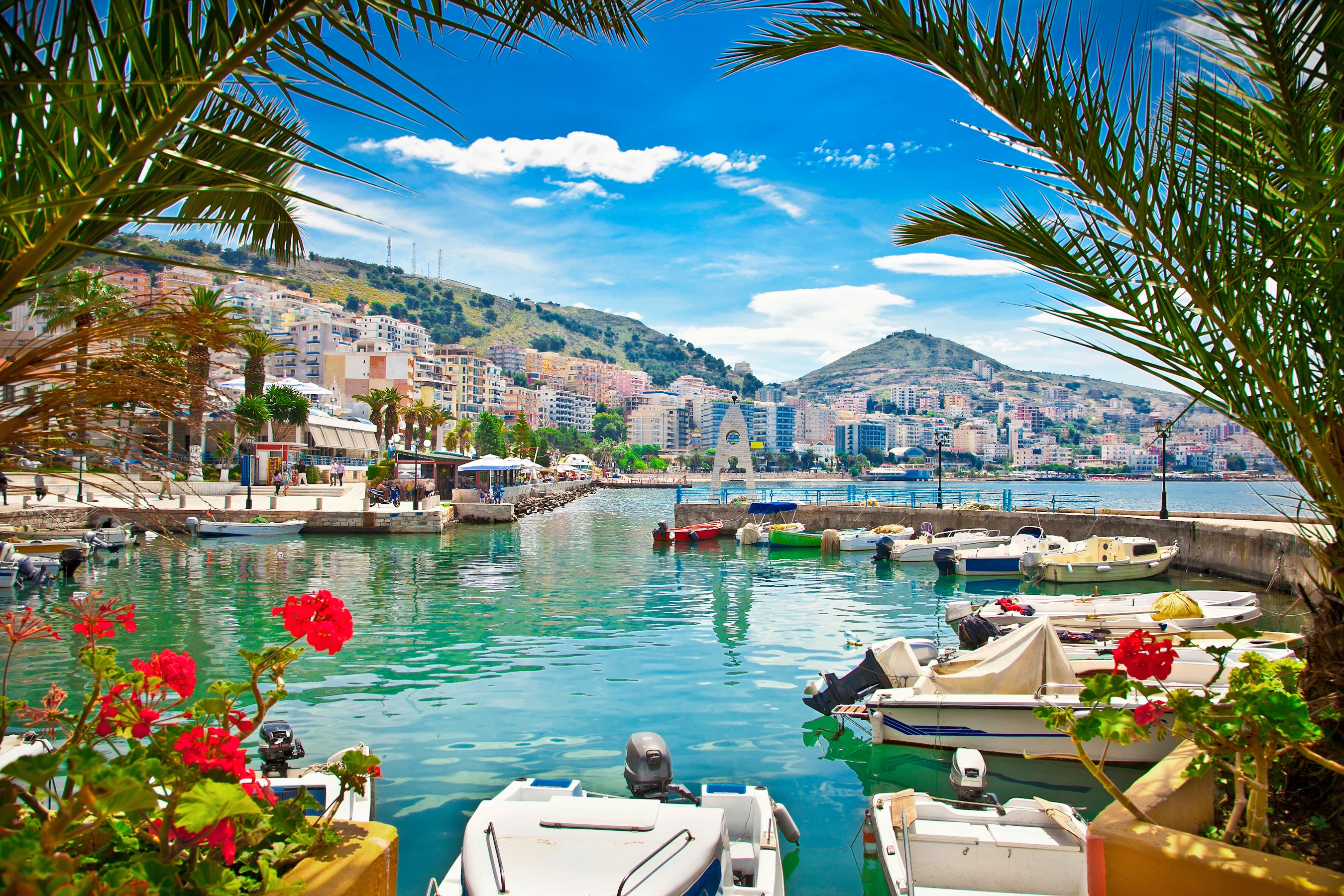 havnen i Saranda med havnepromenaden og husene i bakgrunnen. Motorbåter, palmer og blomster