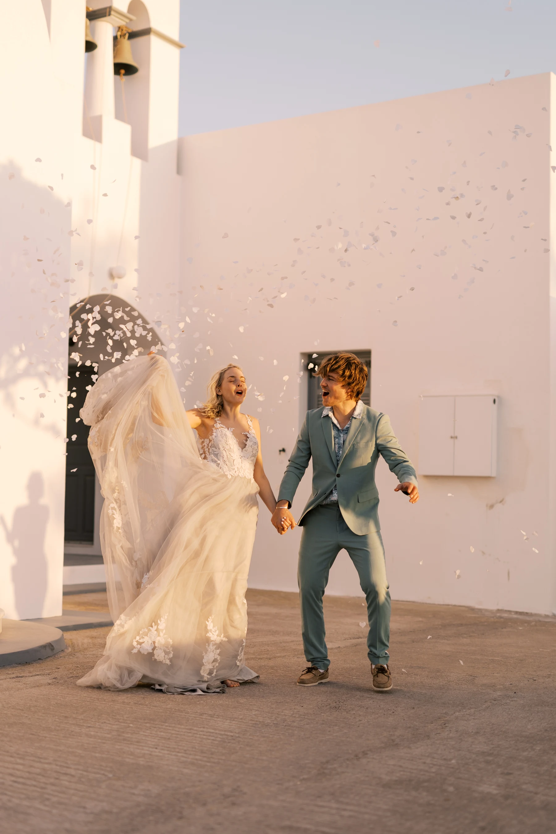 Nygift par feirer i solnedgangen på Santorini. Bruden løfter kjolen og smiler, mens brudgommen i turkis dress ser lykkelig på henne. Hvite blomsterblader svever i luften rundt dem foran en hvit kirke med bjeller.