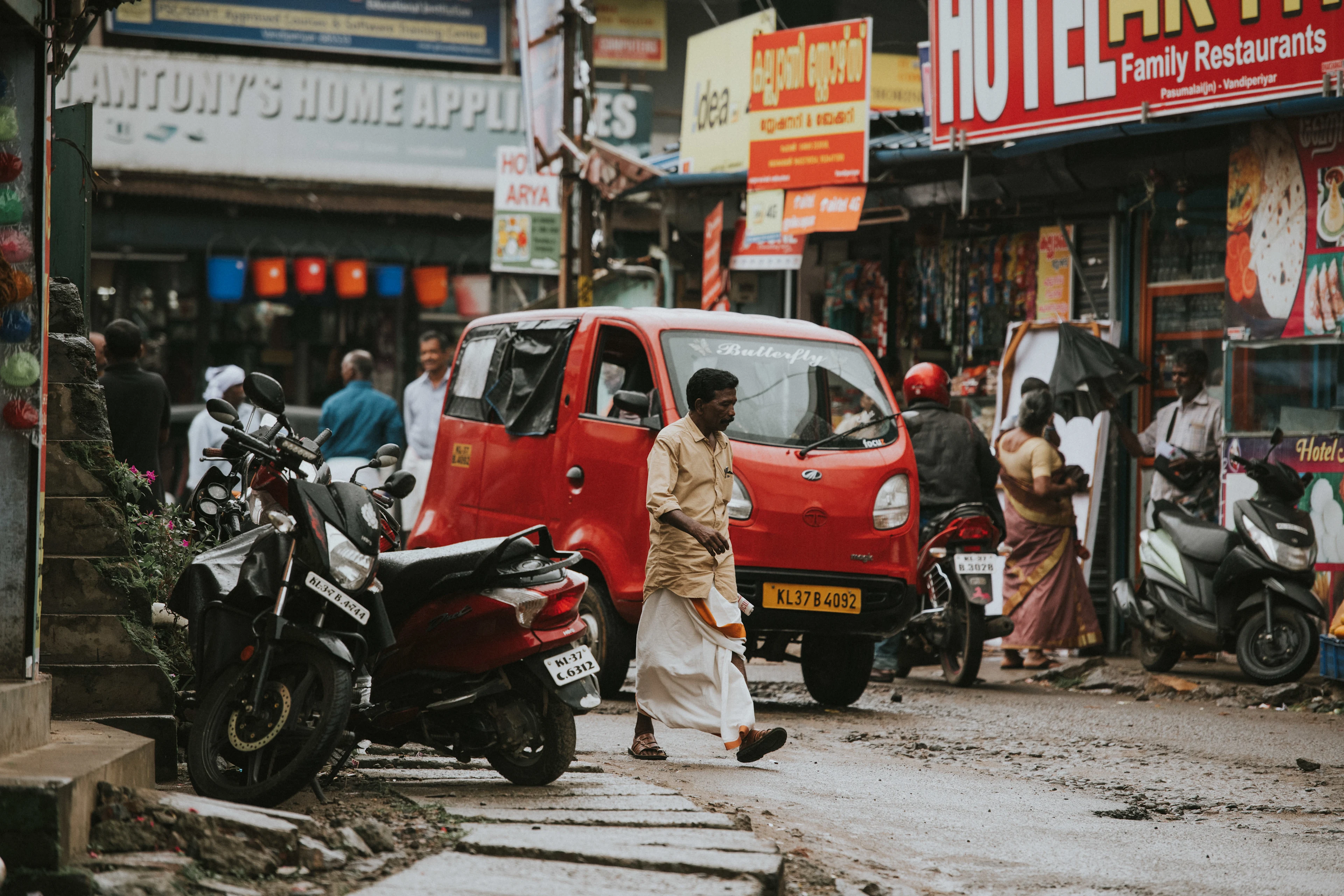 Person som korsar en livlig gata omgiven av motorcyklar, butiker och ett rött fordon i en stadsmiljö