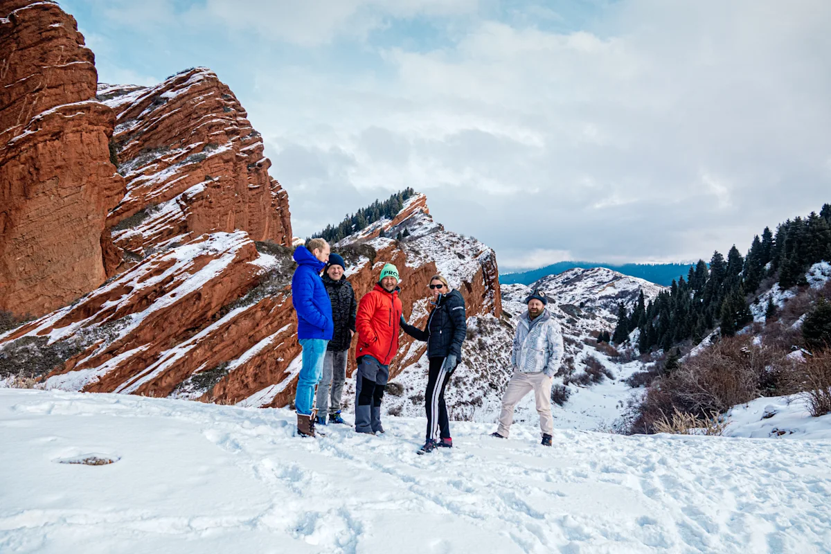 En grupp personer står i snön i ett bergslandskap med röda klippformationer och snöklädda toppar i bakgrunden.