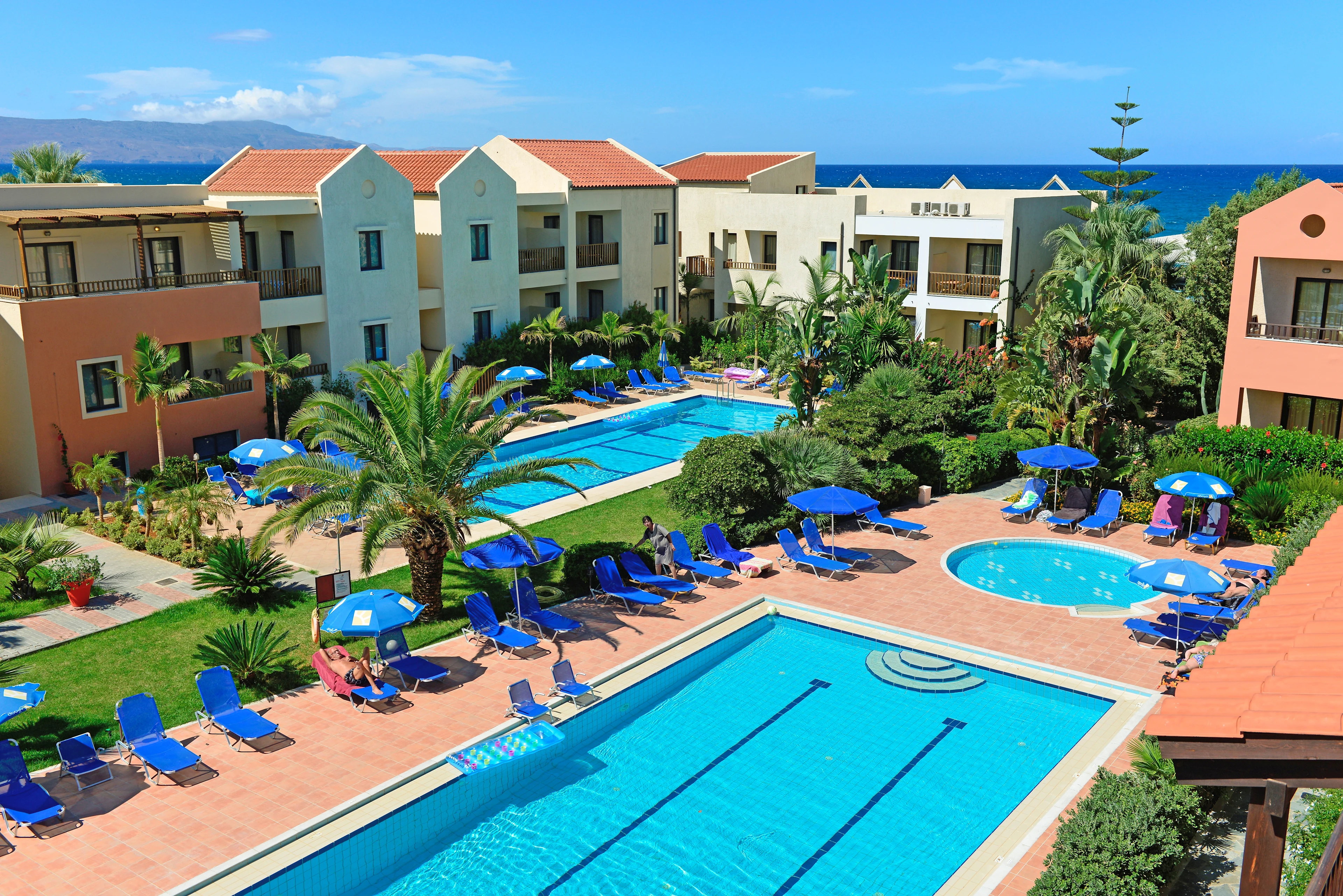 Poolområde med solstolar och grönska på Blue Sea Apartments, med havet i bakgrunden.