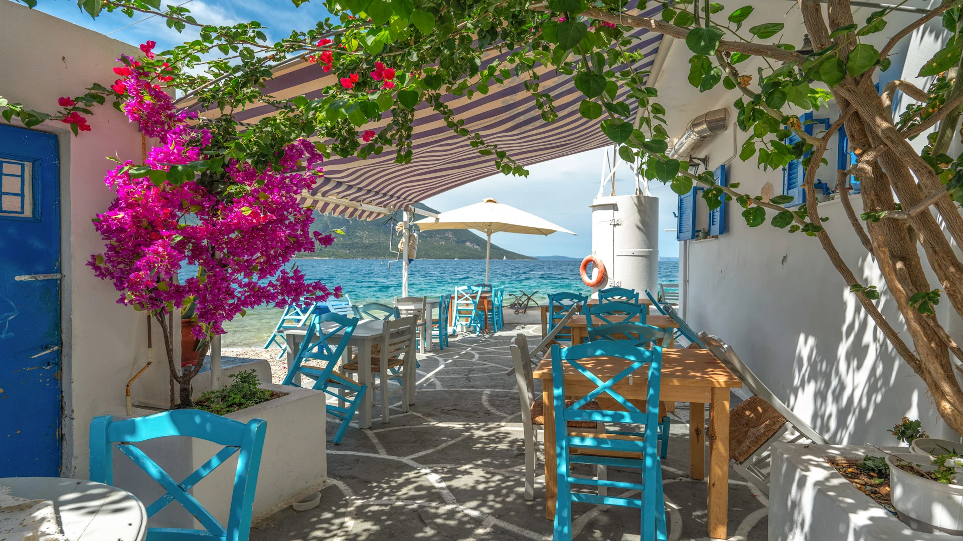 Uteservering vid havet med turkosa stolar, träbord och ett randigt soltak, omgiven av blommande bougainvillea och grönska, med utsikt över klart blått vatten.