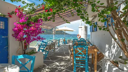 Uteservering vid havet med turkosa stolar, träbord och ett randigt soltak, omgiven av blommande bougainvillea och grönska, med utsikt över klart blått vatten.