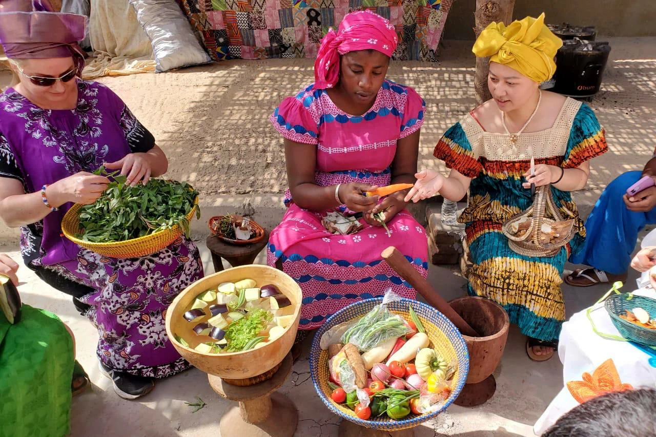 Kolme naista afrikkalaisissa vaatteissa istumassa ja kuorimassa vihanneksia