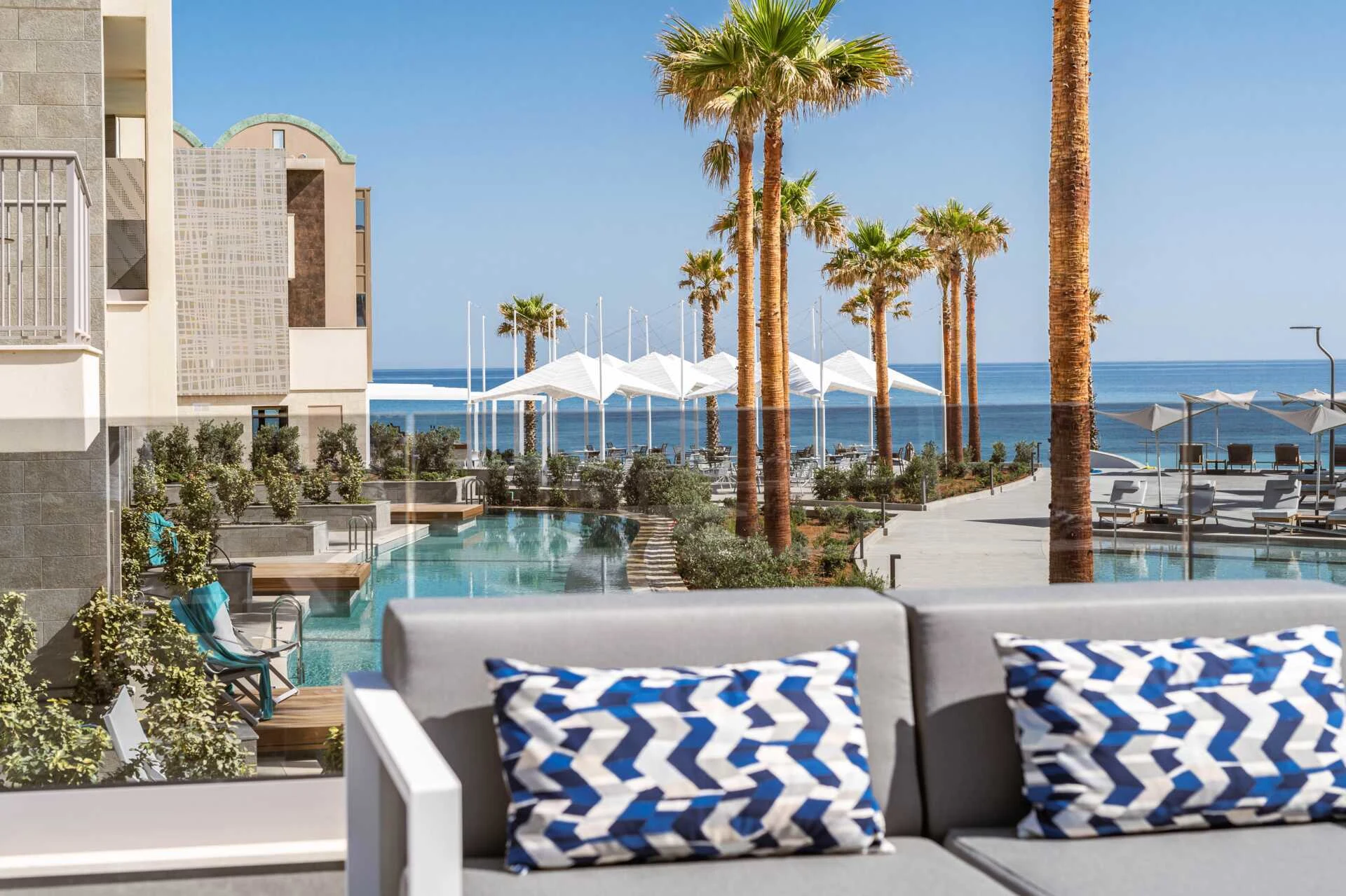 Picture of Amira Luxury Resort on Krete. In the front picture is a sunbed and in the background there is palmtrees, blue sky, and a blue amazing pool. 