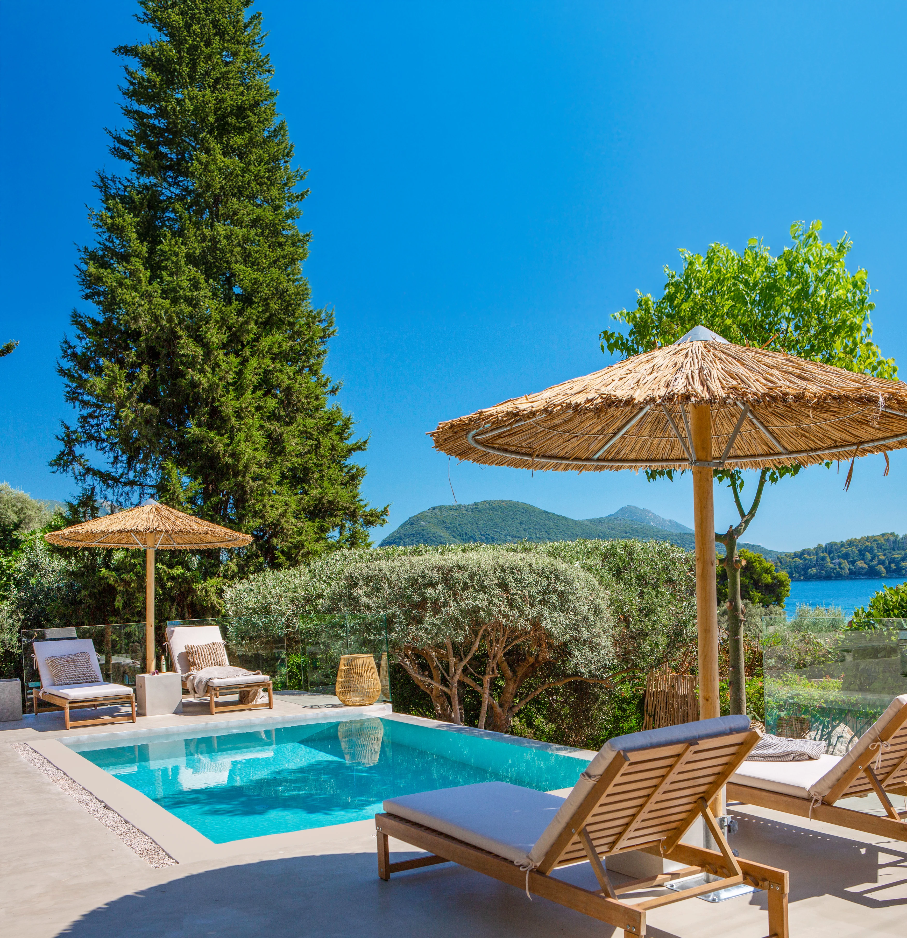 Solig poolterrass med solstolar, parasoller och grönska, med berg och hav i bakgrunden.