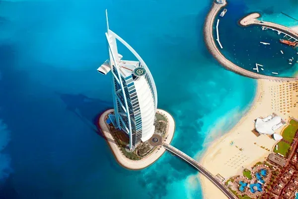 Flygfoto över Burj Al Arab, ett lyxhotell i Dubai med segelformad siluett, beläget på en konstgjord ö omgiven av Persiska vikens blå vatten. En bro förbinder hotellet med fastlandet där sandstränder och byggnader syns.