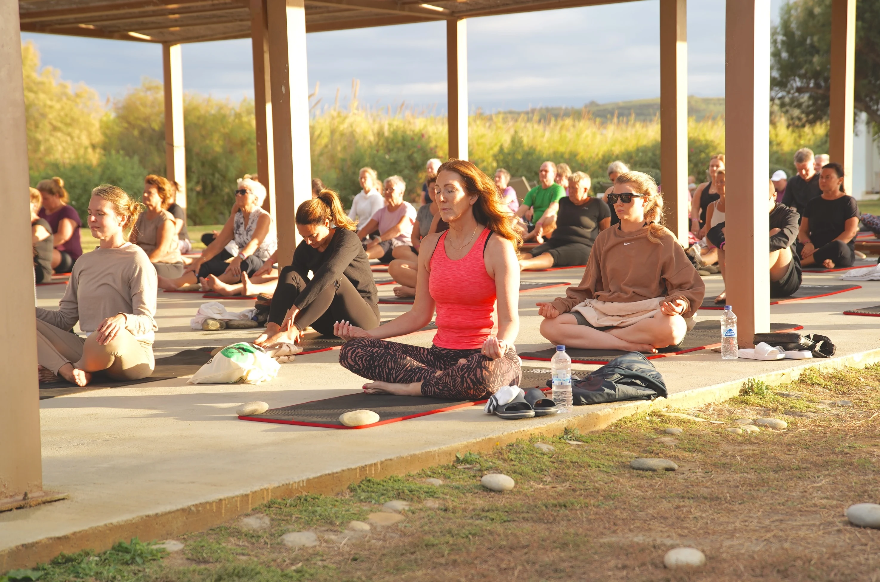 Grupp människor sitter på yogamattor under en öppen paviljong och mediterar utomhus i kvällssol, med grönska och öppet landskap i bakgrunden.