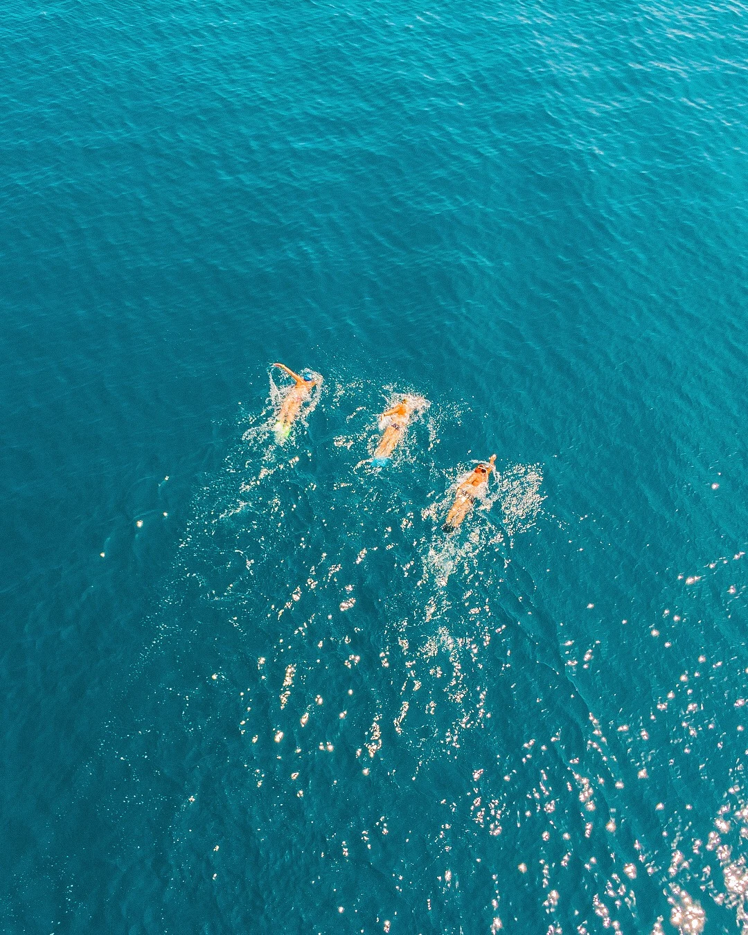  Tre personer simmar i öppet hav, sedda ovanifrån mot ett klart blått vatten. 