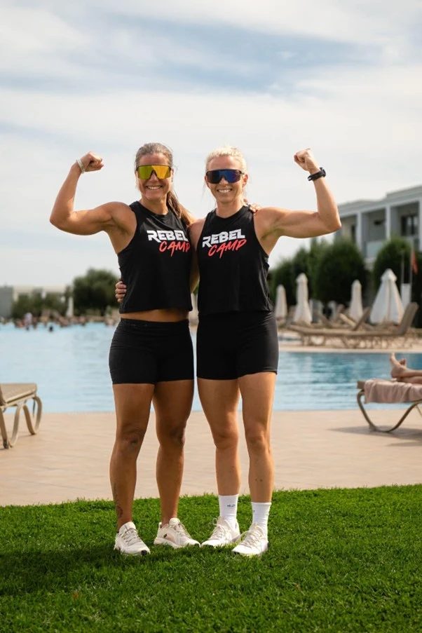 Heidi og Silje står på gresset med et basseng i bakgrunnen. De er begge kledd i sorte treningsshorts og sort treningstopp med påskriften Rebel Camp.  
